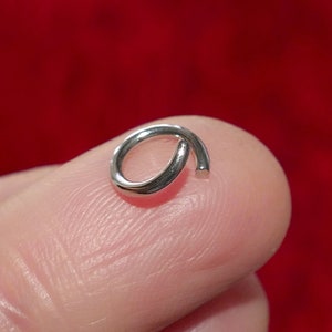 May include: A small, silver-colored open jump ring, likely for jewelry making or crafts. The ring is held on a finger, with a red background. The ring is approximately 0.25 inches in diameter.