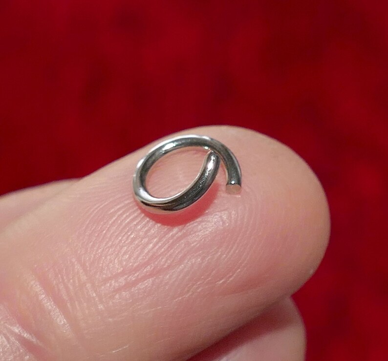 May include: A small, silver-colored open jump ring, likely for jewelry making or crafts. The ring is held on a finger, with a red background. The ring is approximately 0.25 inches in diameter.