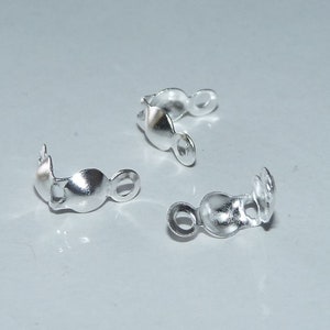 May include: Close-up of several small, silver-colored jewelry findings. Each finding has a curved shape with a loop on one end and a rounded bead-like shape in the middle. The findings are scattered on a light-colored surface.