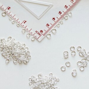 May include: A close-up of a pile of silver jump rings, a ruler, and three smaller piles of jump rings. The jump rings are small, circular metal rings used for connecting jewellery components.