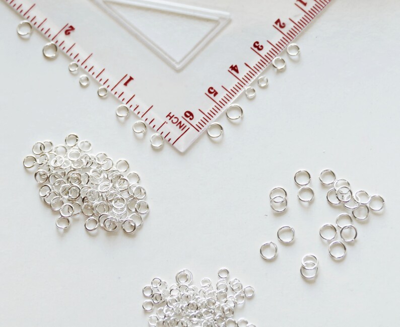 May include: A close-up of a pile of silver jump rings, a ruler, and three smaller piles of jump rings. The jump rings are small, circular metal rings used for connecting jewellery components.
