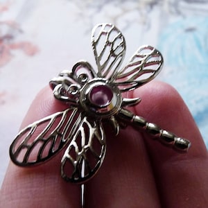 Könnte beinhalten: Silberner Libellen-Anhänger mit einem rosafarbenen Edelstein in der Mitte. Die Flügel der Libelle sind detailliert und filigran.
