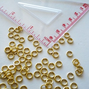 Puede incluir: Una gran cantidad de peque&ntilde;os anillos de salto de color dorado. Los anillos est&aacute;n dispersos sobre una superficie blanca junto a una regla marcada en pulgadas.