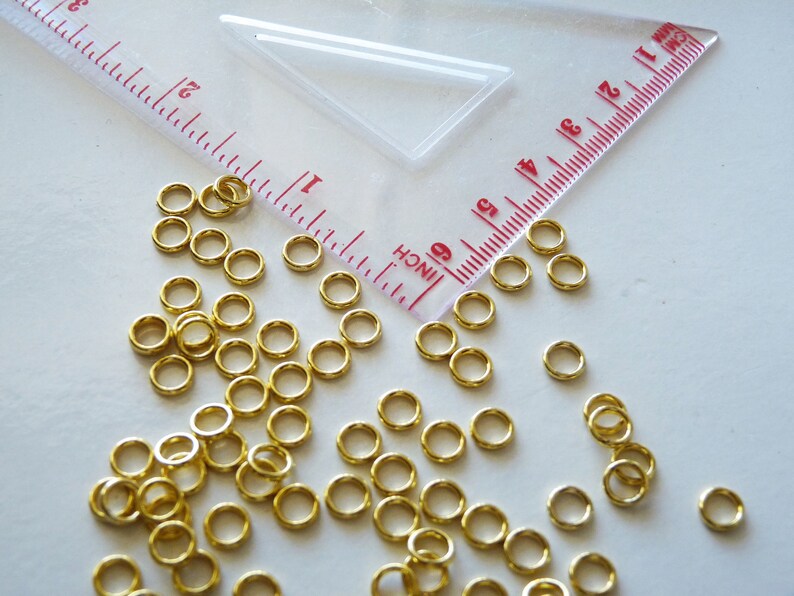 Puede incluir: Una gran cantidad de peque&ntilde;os anillos de salto de color dorado. Los anillos est&aacute;n dispersos sobre una superficie blanca junto a una regla marcada en pulgadas.
