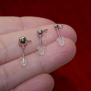 May include: Three silver earring posts with clear rubber stoppers. The earring posts are held in a hand.
