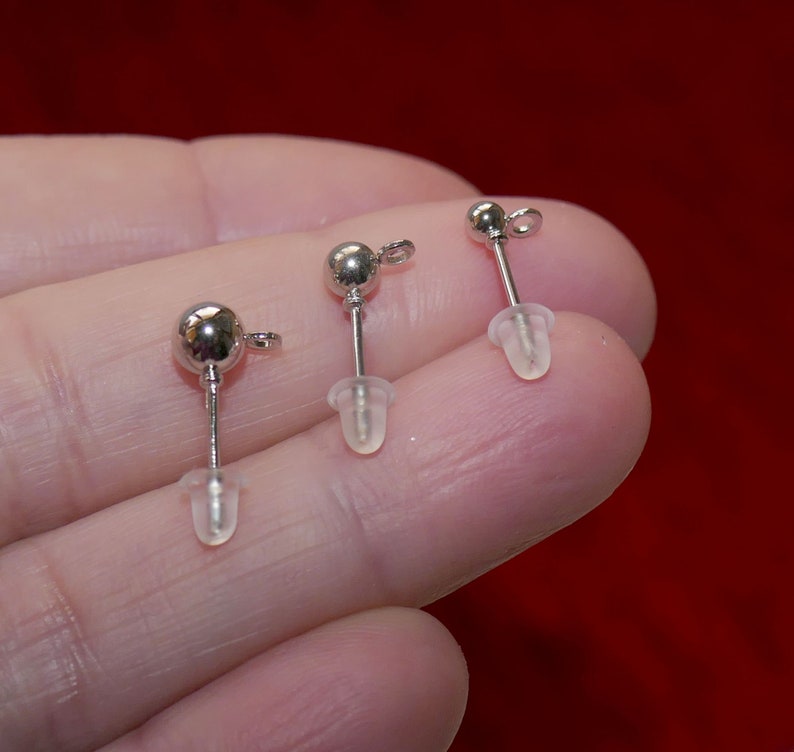 May include: Three silver earring posts with clear rubber stoppers. The earring posts are held in a hand.