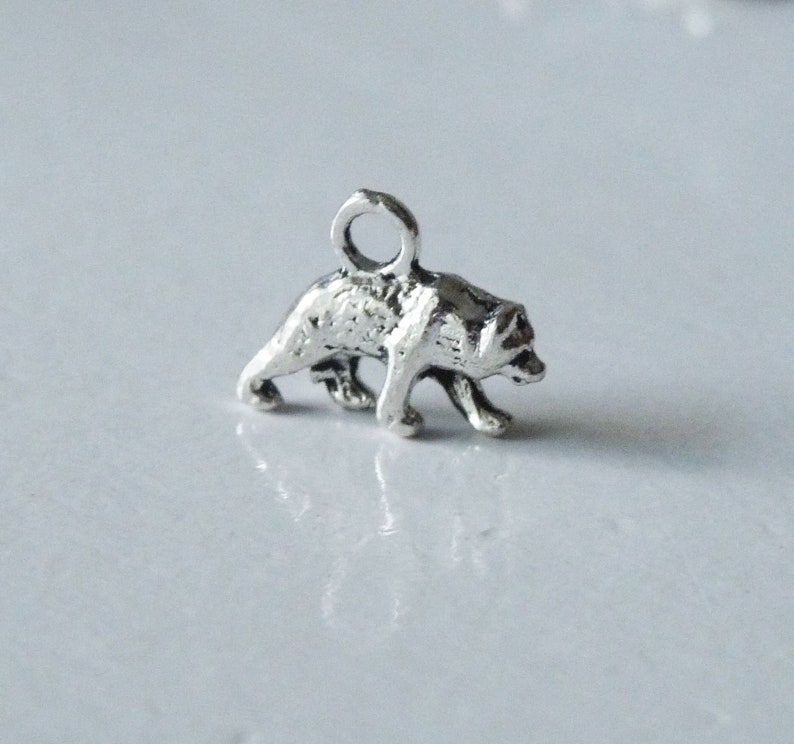May include: Silver charm of a walking bear. The bear is facing right and has a loop at the top for attaching to a necklace or bracelet.