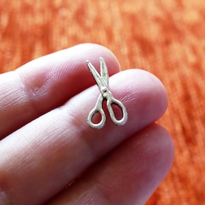 May include: A miniature pair of silver-coloured scissors, held in a person's hand.