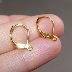 Puede incluir: Dos ganchos para pendientes de color dorado, también conocidos como cierres de palanca para pendientes, se muestran en un dedo. Los ganchos son de metal y tienen un acabado liso y pulido.