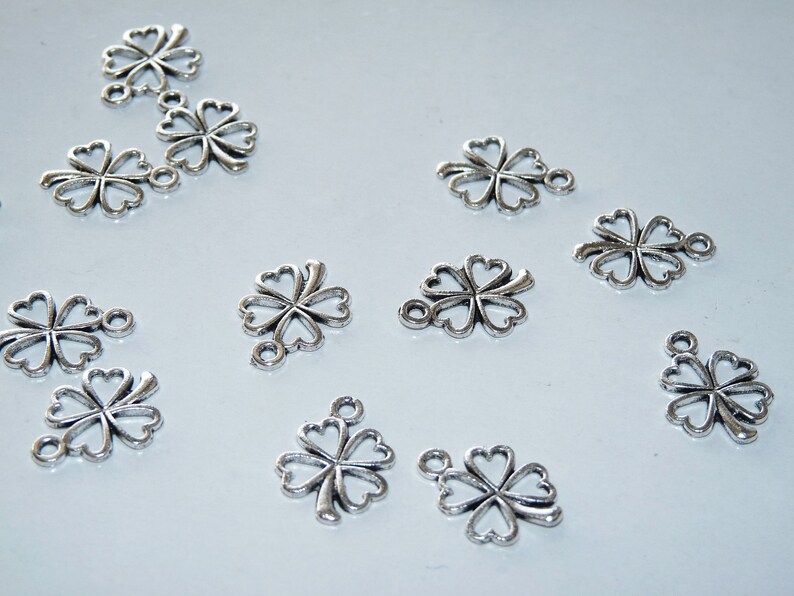 May include: A collection of silver-toned metal charms shaped like four-leaf clovers. Each charm has a loop at the top for attaching to jewelry or other crafts.