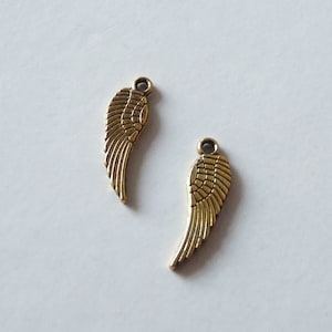 Könnte beinhalten: Zwei goldfarbene Metall-Engel Flügel Charms. Die Flügel sind mit einem strukturierten Muster verziert und haben eine Schlaufe oben zum Anbringen an Schmuck oder anderen Bastelarbeiten.
