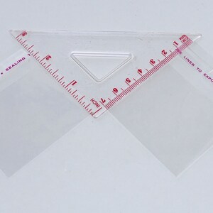 Puede incluir: Una regla de pl&aacute;stico transparente con marcas rojas para medir en pulgadas. La regla tiene dos tri&aacute;ngulos con el texto "OKER BRAND SEALING" y "LINER TO EXPOSE ADHESIVE" impreso en ellos.