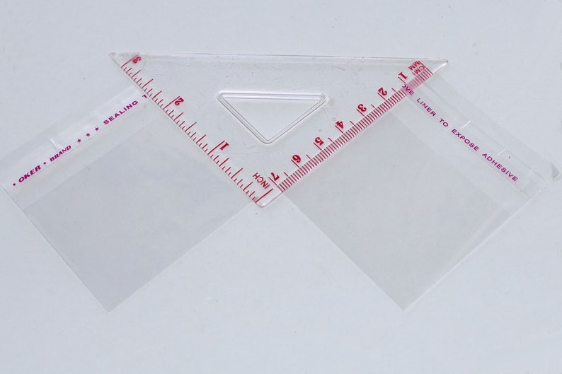 Puede incluir: Una regla de pl&aacute;stico transparente con marcas rojas para medir en pulgadas. La regla tiene dos tri&aacute;ngulos con el texto "OKER BRAND SEALING" y "LINER TO EXPOSE ADHESIVE" impreso en ellos.
