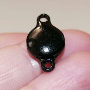 Puede incluir: Encanto de metal negro con una superficie redonda y brillante y dos bucles. El encanto tiene un diámetro de aproximadamente 2,5 cm.