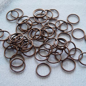 Puede incluir: Una colección de pequeños anillos de salto de color bronce dispersos sobre una superficie clara. Los anillos son circulares y parecen ser de metal, adecuados para la fabricación de joyas o proyectos de artesanía.
