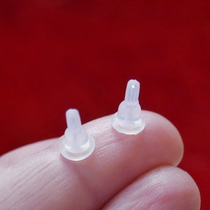 May include: Two small, translucent silicone bottle nipples with a rounded base and a narrow, ridged tip. The nipples are held against a red background, showcasing their delicate design and form.