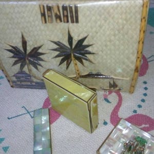 May include: Vintage woven handbag with bamboo handles and the word "HAWAII" on the front. The bag features a palm tree design. Also included are a small yellow compact, a rectangular mother-of-pearl piece, and a square compact with a decorative emblem.