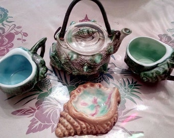 Könnte beinhalten: Eine grüne und braune Teekanne aus Keramik mit Muschel-Design, zwei passende Tassen und eine schalenförmige Schale. Die Teekanne hat einen Deckel mit einem grünen Schildkröten-Design. Die Tassen und die Schale sind ebenfalls mit Muschel-Designs verziert.