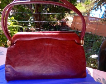 Può includere: Una borsa a mano in pelle bordeaux con una chiusura dorata e un manico superiore. La borsa ha una superficie testurizzata e squamosa.