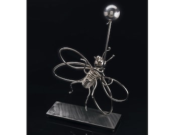 Peut inclure: Une sculpture en métal argenté représentant une abeille avec des ailes en fil de fer et un corps en spirale, montée sur une base rectangulaire. Une longue antenne fine s'étend vers le haut, surmontée d'une sphère argentée. La sculpture est exposée sur fond noir.