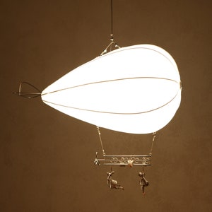 Puede incluir: Una lámpara blanca en forma de lágrima cuelga del techo. La lámpara está hecha de metal y tela y se asemeja a un globo aerostático. Dos pequeñas figuras decorativas cuelgan de la parte inferior de la lámpara.