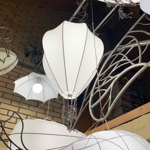 Puede incluir: Pantallas de lámparas blancas de tela con forma de globos aerostáticos y dirigibles colgando de un marco de metal. Las lámparas están encendidas y emiten un brillo cálido.