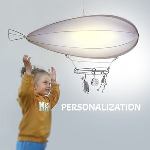 May include: A white, round, hanging light fixture shaped like a blimp with a metal frame and hanging figures. The light is on and illuminating the room. A young person is standing in front of the light with their arms raised. The text "PERSONALIZATION" is printed below the light.