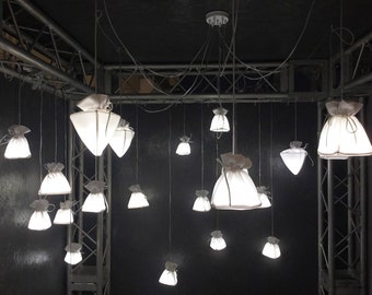 May include: A collection of white fabric lampshades hanging from a metal frame. The lampshades are shaped like pouches or bags and are illuminated from within, casting a soft, warm glow.