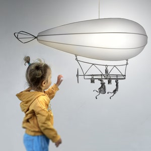 Puede incluir: Un niño pequeño extiende la mano hacia un dirigible blanco colgante con un marco de metal y figuras esqueléticas colgando de él.