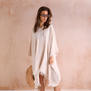 Puede incluir: Un poncho de algod&oacute;n blanco con detalles de flecos y capucha con cord&oacute;n. El poncho lo lleva puesto una persona que lleva gafas de sol y sandalias.