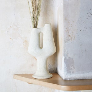 May include: A cream-colored, abstract ceramic vase with a unique cutout design, holding dried ornamental grasses and seed pods. The vase sits on a wooden shelf against a textured, off-white wall. The overall aesthetic is minimalist and natural.