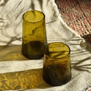 Peut inclure: Deux verres à boire en verre vert olive sur un fond de tissu en lin blanc. Les verres sont en verre épais et ont une forme arrondie.