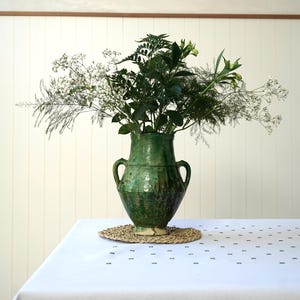 May include: A green glazed ceramic vase with two side handles, featuring a mottled finish and a tapered base. The vase is filled with a lush arrangement of green foliage, delicate white baby's breath, and slender stems of greenery.