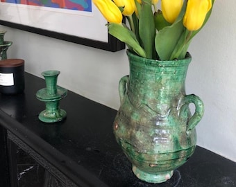 Könnte beinhalten: Ein Strauß gelber Tulpen in einer grünen Keramikvase mit zwei Henkeln. Die Vase hat ein rustikales, gealtertes Aussehen. Die Tulpen sind einfach und elegant angeordnet.
