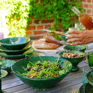 May include: A collection of dark green ceramic tableware including a large salad bowl filled with mixed greens, smaller bowls, and plates. The pieces feature a glossy, textured finish with scalloped edges, arranged on a wooden surface.
