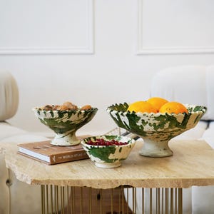 Op de afbeelding: Drie keramische kommen met groen-witte spetspatronen staan op een stenen tafel. De kommen zijn gevuld met walnoten, sinaasappels en rode bessen. Een boek met de titel "The World Atlas" ligt ook op de tafel.