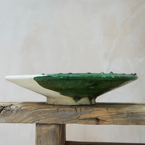 Puede incluir: Un cuenco de cer&aacute;mica hecho a mano con esmalte blanco y verde. El cuenco tiene una forma ancha y poco profunda con un borde ligeramente elevado. El esmalte verde cubre la mitad superior, creando un contraste con la base blanca. El cuenco se exhibe sobre una superficie de madera r&uacute;stica.