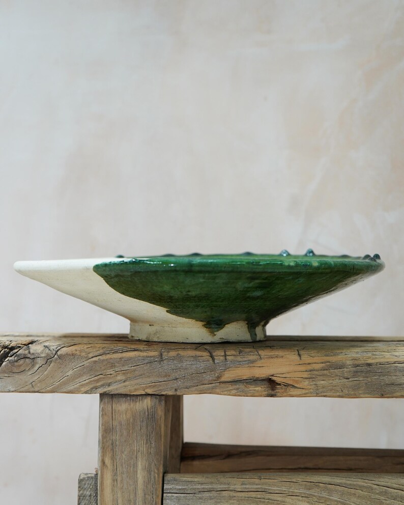 Puede incluir: Un cuenco de cer&aacute;mica hecho a mano con esmalte blanco y verde. El cuenco tiene una forma ancha y poco profunda con un borde ligeramente elevado. El esmalte verde cubre la mitad superior, creando un contraste con la base blanca. El cuenco se exhibe sobre una superficie de madera r&uacute;stica.