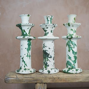 May include: Three white ceramic candle holders with green splatter paint design. The candle holders are of varying heights and sit on a weathered wooden bench. The candle holders have a classic, elegant shape.