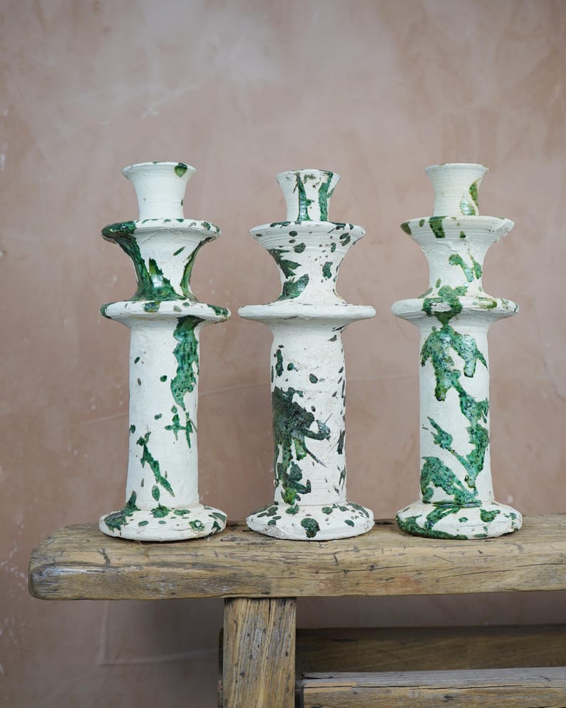 May include: Three white ceramic candle holders with green splatter paint design. The candle holders are of varying heights and sit on a weathered wooden bench. The candle holders have a classic, elegant shape.
