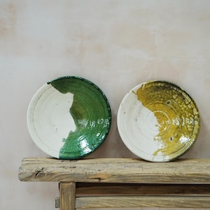 Puede incluir: Dos platos de cer&aacute;mica con un dise&ntilde;o r&uacute;stico. Uno es blanco y verde, el otro blanco y amarillo. Los platos est&aacute;n sobre un banco de madera envejecida contra una pared de color neutro. Los platos tienen un aspecto &uacute;nico y artesanal.