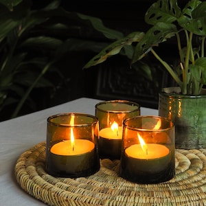 May include: Three green glass candle holders with lit candles on a woven straw placemat. The candles are burning brightly and casting a warm glow.