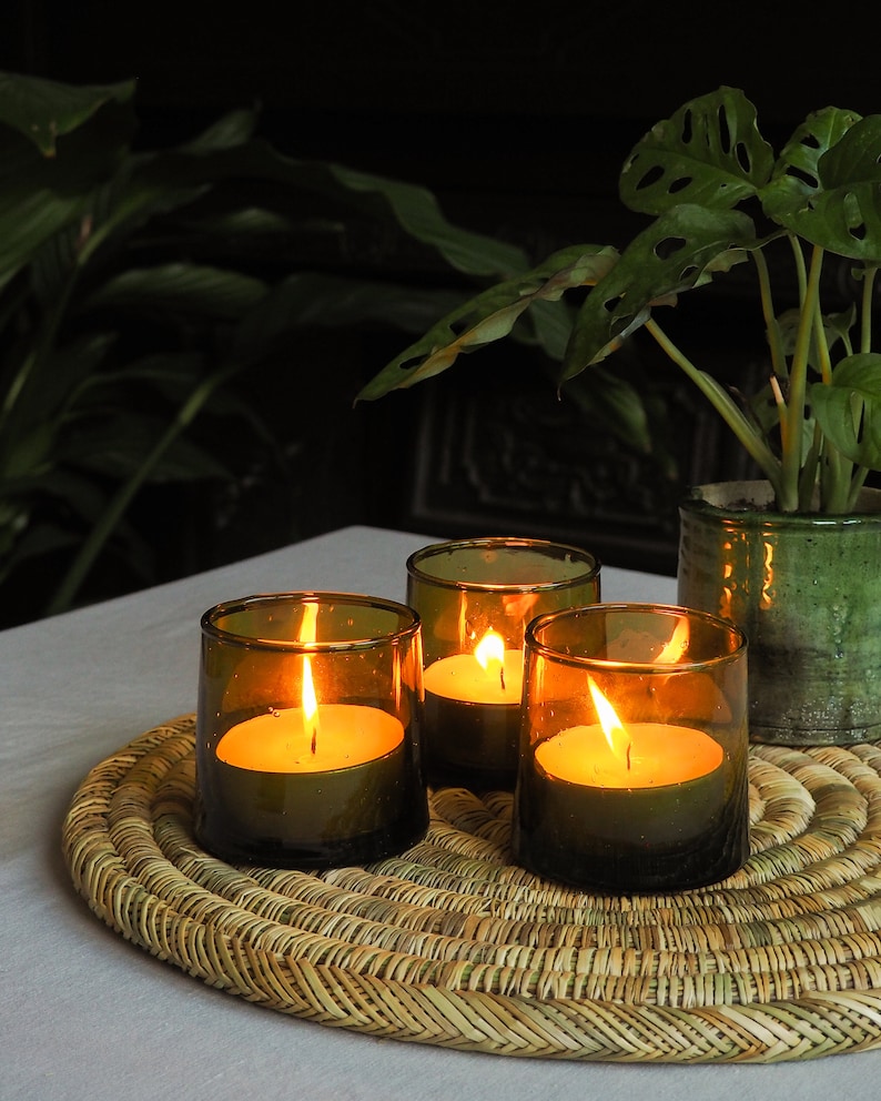 May include: Three green glass candle holders with lit candles on a woven straw placemat. The candles are burning brightly and casting a warm glow.