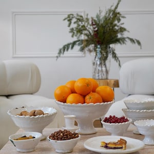May include: A collection of white ceramic footed bowls and plates with scalloped edges. The bowls are filled with fresh oranges, walnuts, hazelnuts, and red currants. A small plate holds a slice of fruit tart, creating a rustic table display.