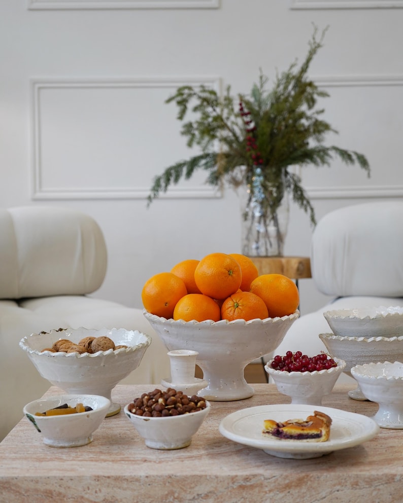 May include: A collection of white ceramic footed bowls and plates with scalloped edges. The bowls are filled with fresh oranges, walnuts, hazelnuts, and red currants. A small plate holds a slice of fruit tart, creating a rustic table display.