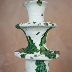 May include: A handcrafted ceramic candlestick with a white base and vibrant green glaze accents. The candlestick features a tiered design with a flared top and base, showcasing a unique, artistic aesthetic. The textured surface adds to its artisanal charm.