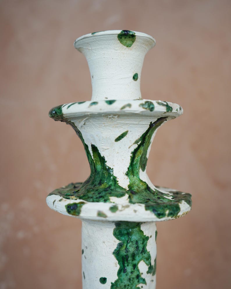 May include: A handcrafted ceramic candlestick with a white base and vibrant green glaze accents. The candlestick features a tiered design with a flared top and base, showcasing a unique, artistic aesthetic. The textured surface adds to its artisanal charm.