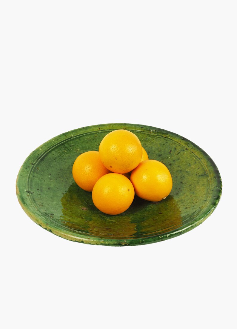 May include: A rustic, green ceramic plate holds five bright orange oranges. The plate has a slightly uneven, aged appearance, with a textured surface. The oranges are round and vibrant, contrasting with the plate's color.