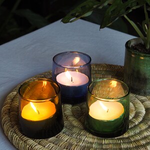 May include: Three candles burning in glass jars on a woven wicker placemat. The candles are white and are in blue, green, and brown glass jars.