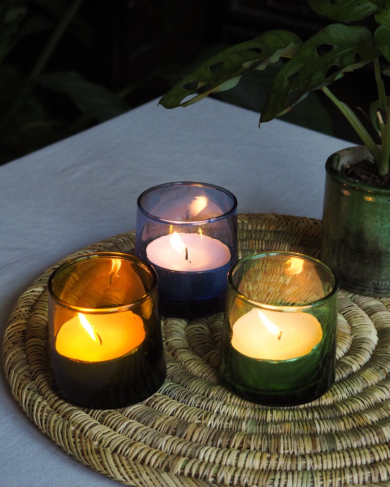 May include: Three candles burning in glass jars on a woven wicker placemat. The candles are white and are in blue, green, and brown glass jars.
